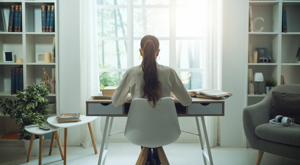 Kvinne sitter ved skrivebord i sin egen stue. Symmetrisk.