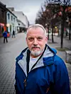 Eldre mann i blå jakke står på en gågate med bygårder i bakgrunnen