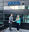 To personer håndhilser foran MDCO-skiltet på en mørk glassfasade.