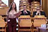 Agnes Viljugrein står i Stortinget med papirer mens to menn sitter bak henne under monarkidebatten.