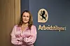 Kvinne i rosa skjorte står foran skilt med Arbeidstilsynet-logo og navn.