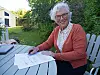 Eldre kvinne med briller sitter ved et hagebord og leser dokumenter.
