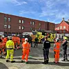 Brannmannskaper samlet foran en gul og rød brannbil mellom bygårder.