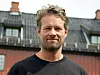 Smilende mann i mørk genser foran en eldre bygning med skifertak.