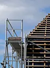 Stillas ved siden av en stor trekonstruksjon med synlige taksperrer mot en skyet himmel.