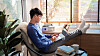 Ung asiatisk mann bruker laptop i en lys stue med planter og persienner.