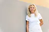 Blond kvinne i hvite klær står foran en lys grå vegg.