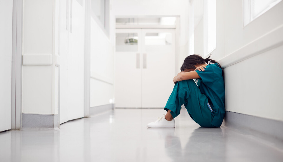 Kvinnelig sykepleier sitter med hodet i hendene i en sykehuskorridor.