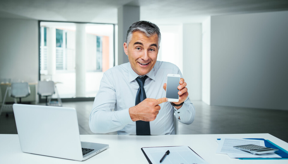 Mann sitter smilende ved kontorpult og peker på mobiltelefonen sin.