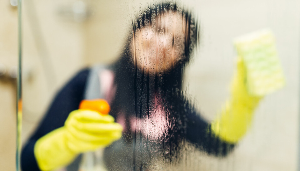 Kvinnelig renholdsarbeider som vasker et bad, fotografert gjennom en våt dusjvegg.
