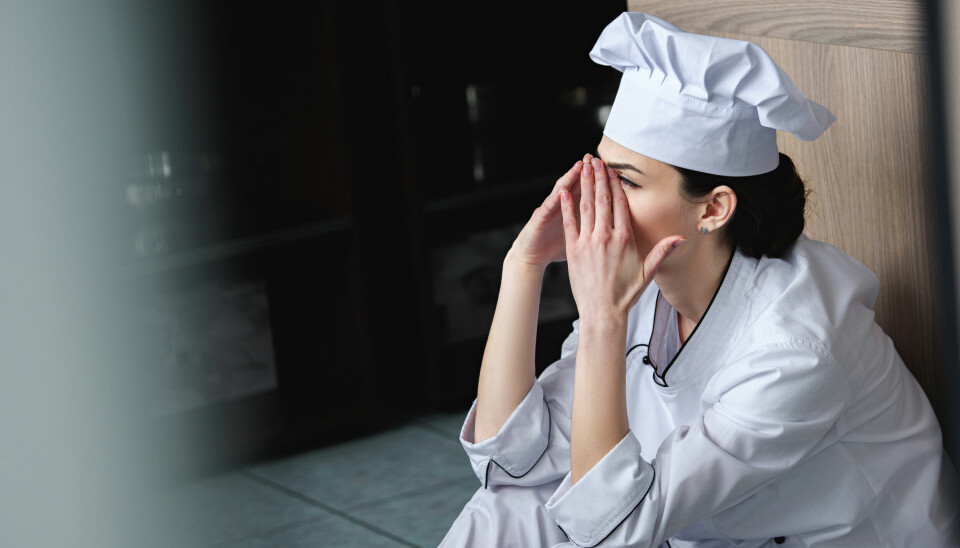 Ung kvinne i kokkeklær sitter på kjøkkengulv med ansiktet i hendene.