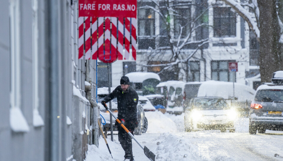 Mann rydder snø på et fortau. Skilt med «fare for ras» på husveggen.